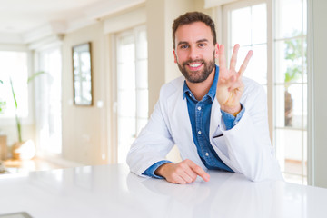 Handsome doctor man wearing medical coat at the clinic showing and pointing up with fingers number three while smiling confident and happy.