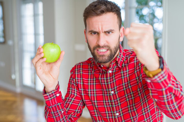 Handsome man eating fresh healthy green apple annoyed and frustrated shouting with anger, crazy and yelling with raised hand, anger concept