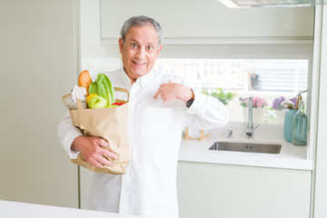 Handsome senior man holding a paper bag of fresh groceries at the kitchen with surprise face pointing finger to himself