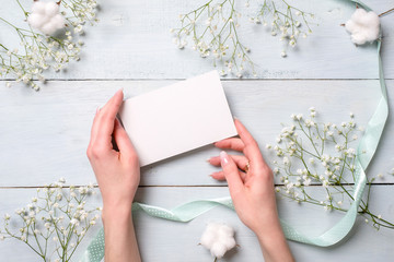 Hands holding blank paper card on light blue wooden desk with flowers. Tender greeting card for womens or mothers day, easter, spring holidays.