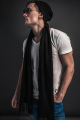 Young men model tests in studio.  Fashion shooting beautiful guy in blue jeans, white shirt, black scarf, hat and glasses. Posing on a gray background.