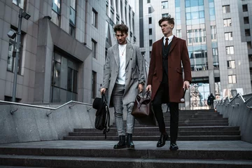 Two fashion men models posing on city street. Wearing in classic and casual closes. Suit, coat, shirt, sweater, boots and leather bag. Men model test near business center © Yevhen