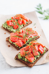 Close-up view of three sandwiches with rye bread, avocado and smoked salmon on a white kitchen table.