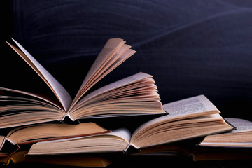 Open books are a stack on the desk, against the background of a chalk board. Difficult homework at school, a mountain of knowledge.