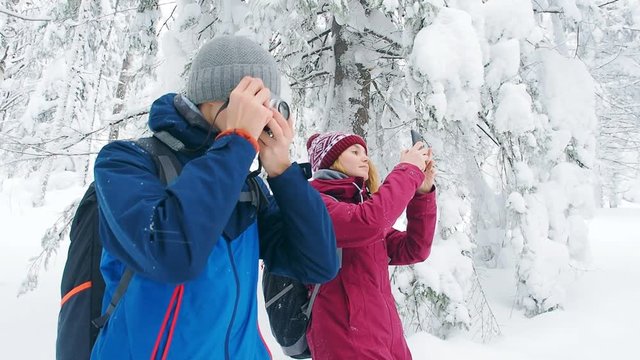 Young couple hiking taking photo outdoors winter landscape snow forest. Happy young man with camera takes a photo of snow trees and hiking girl taking picture by smartphone.