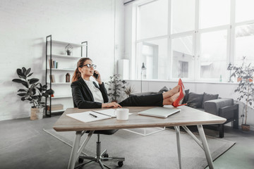 Pretty young woman sits in chair and holds legs on table. She wears black suit and red high heels. Girl talks on the phone. She is busy