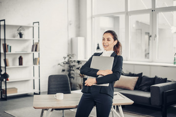 Cheerful and amazing girl stands and sniles. She holds laptop in hands. She is laughing. Girl wears black suit. She is in white office room