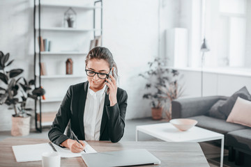 Serious and concentrated girl sits at table and works. She talks on phone and writes down information. She is in white office room with green plants