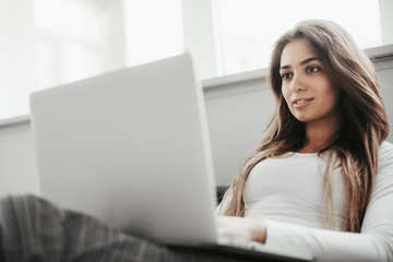 Fototapeta premium Portrait of busy girl looks on laptop she holds on knees. Woman is leaning to white wall. She is concentrated