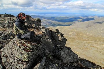 Caucasian white male tourist in sportswear, buff and glasses sits on a rock against the backdrop of mountains