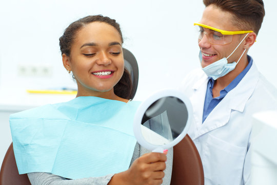 Cute Woman Looking At Mirror In Hand While Doctor Standing Near.