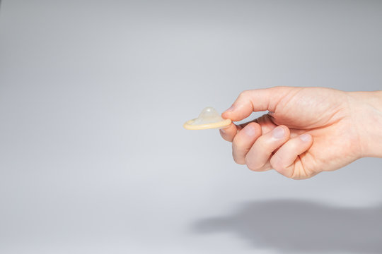 Man's hand holding an unused and rolled-up condom between thumb and index finger, light gray background. Concept photo of contraception and safe sex. Copy space.