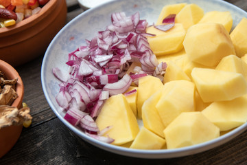 Sliced potato and onion on ceramic plate, and other vegetables, spices and dried mushrooms in the background. Old wooden table.