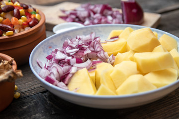 Sliced potato and onion on ceramic plate, and other vegetables, spices and dried mushrooms in the background. Old wooden table.