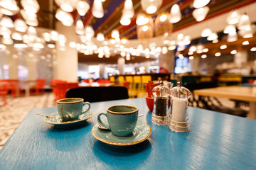 A cup of coffee on a saucer with a spoon stands on a blue table. Cup in focus. The background of the cafe is blurred.