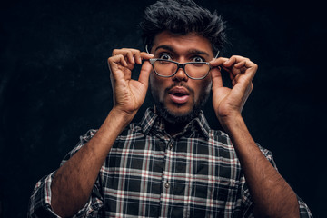 Surprised Indian guy lowers his glasses and looks at the camera. Studio photo against a dark textured wall