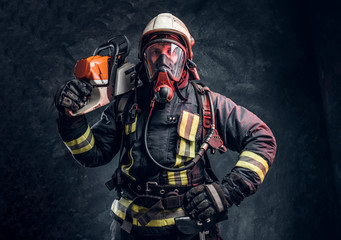 Naklejka premium A brave firefighter wearing full protective equipment posing with a chainsaw on his shoulder. Studio photo against a dark textured wall