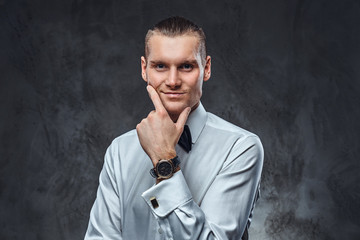 Confident elegantly dressed man wearing white shirt and bow tie, touching his face and looking at a camera. Studio shot on a dark textured wall