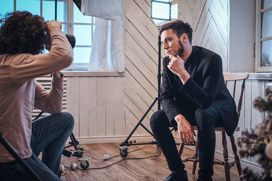 Professional Photo Shooting At The Studio: A Handsome Young Guy Sitting, Posing And Holds Arm Under His Chin The Curly Photographer Is Taking Pictures With A Digital Camera 