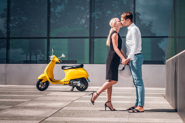 Young couple in love, hold hands and kiss standing under a skyscraper on hot summer day
