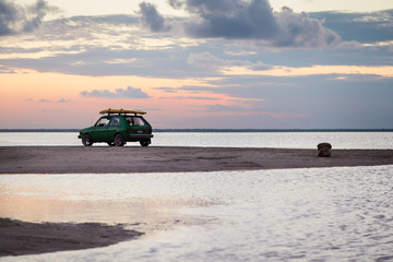 Fototapeta na wymiar A retro surf car with a surfboard on a sand bar near the ocean with the sun setting in the background.