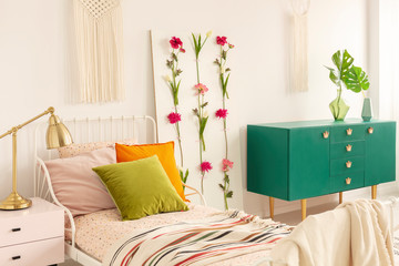 Flower board between single bed with olive green, orange and pastel pink pillows and green wooden cabinet with leaf in glass vase