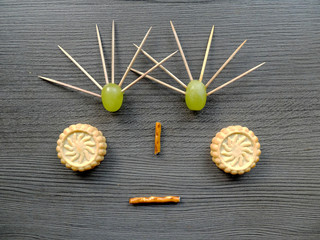 Funny face made of food (grapes and salty sticks). Neutral expression, long eyelashes, small mouth.