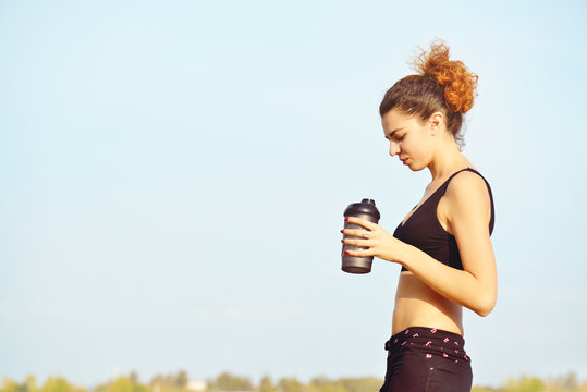 Beautiful healthy woman drinking water against blue background. Side view of girl in sport clothes with bottle.