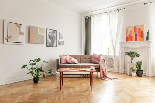 Wooden table in front of sofa with pink blanket in apartment interior with posters and plants. Real photo