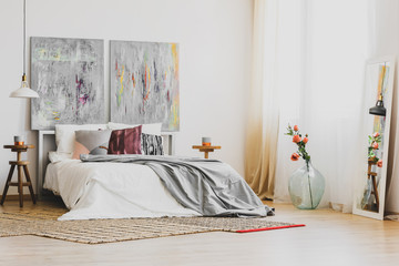 Natural rug on wooden floor of stylish bedroom interior with king size bed, wooden nightstands and grey fancy paintings on white wall