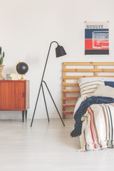 Real photo of a modern lamp in a retro bedroom interior with a bed, poster and wooden cupboard