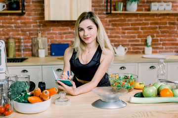 Young pretty woman in the kitchen at home writing shopping list and watching at camera. Modern kitchen, fresh vegetables and healthy food on the wooden table.