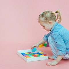 A charming little girl in denim clothes on a pink background puts wooden multi-colored cubes in a white box. Copy space.