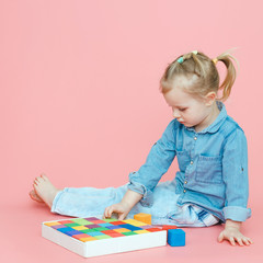 A charming little girl in denim clothes on a pink background puts wooden multi-colored cubes in a...