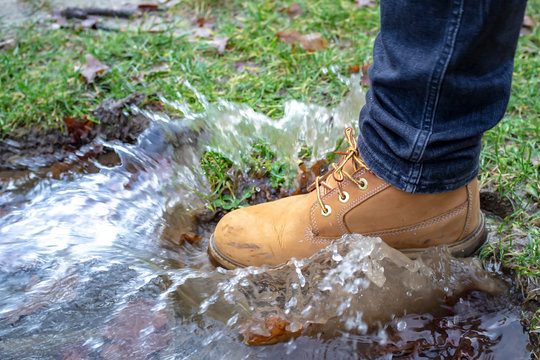 Man's leg in dirty, yellow boots confidently walks into a puddle and splashes of water scatter in different directions, spring day in the countryside.