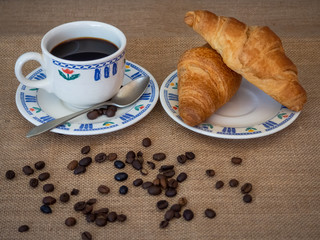 Breakfast of a porcelain coffee cup, an antique spoon, two croissant and coffee beans on a vintage burlap background