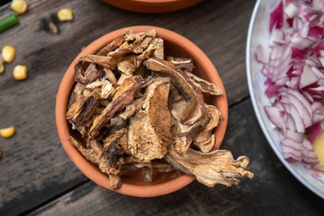 Dried boletus mushrooms in rustic ceramic bowl on old wooden table, and other mixture of vegetables and spices in the background. 