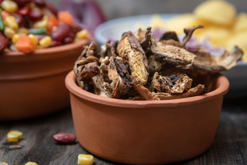 Dried boletus mushrooms in rustic ceramic bowl on old wooden table, and other mixture of vegetables and spices in the background. 