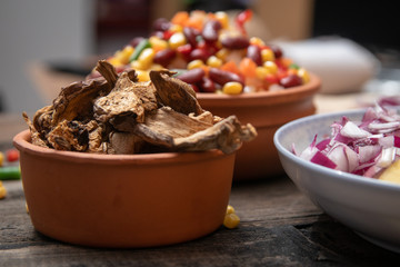 Dried boletus mushrooms in rustic ceramic bowl on old wooden table, and other mixture of vegetables and spices in the background. 