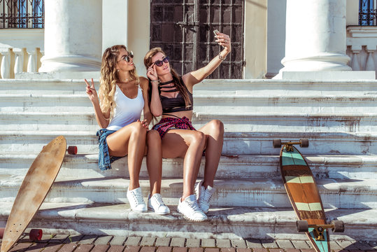 Two girls girlfriends girlfriends, sit on steps, skateboard longboards, take pictures on phone, online video call social networks, selfie. Women pass on helper, happy smiling rest in summer in city.