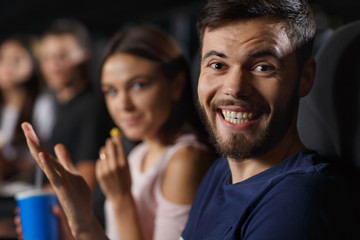 Closeup of positive man with big smile looking at camera at cinema theater. Young bearded man enjoying movie review and laughing at funny comedy. Concept of entertainment and happiness.