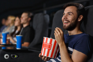 View from side of excited man with big smile laughing at funny comedy at cinema. Bearded young man eating tasty popcorn and enjoying interesting film. Concept of entertainment and leisure.