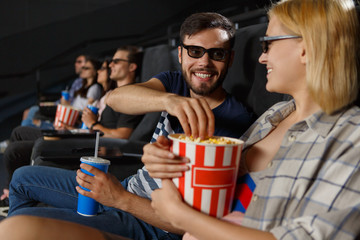 Cute couple having romantic date at cinema. Attractive girl and bearded man in 2d glasses, laughing, eating popcorn, drinking sparkling water and enjoying new film. Concept pf leisure and happiness.