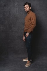 Handsome young man on gray background looking at camera. Portrait of young man with hands in pockets leaning against gray wall.