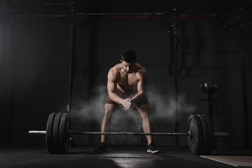 Obraz na płótnie Canvas Crossfit athlete clapping hands and preparing for weight lifting at the gym. Barbell magnesia protection dust cloud. Handsome man doing functional training. Practicing powerlifting. Workout exercises.