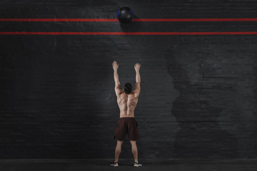 Crossfit athlete throws ball at the gym. Handsome man doing functional training. Workout exercises. Brick wall copy space. Place for text.