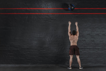 Crossfit athlete throws ball at the gym. Handsome man doing functional training. Workout exercises. Brick wall copy space. Place for text.