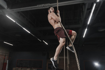 Young crossfit athlete climbing a rope at the gym. Handsome man doing functional training. Workout exercises