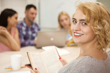 Positive and pretty blonde female student sitting smiling at light class. Young girl wearing in gray t-shirt, with bright lipstick, holding book in hands at university and turning at camera.