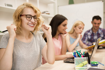 Happy blonde girl in glasses gesturing by hands and smiling and while sitting on lesson. Group of students sitting at table, studying and doing homework using notes and laptop in classroom.